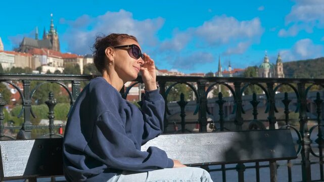 Tourist enjoying prague castle view from river bank. Young woman wearing sunglasses and casual clothes sits on a bench, enjoying the panoramic view of prague castle from the bank of vltava river