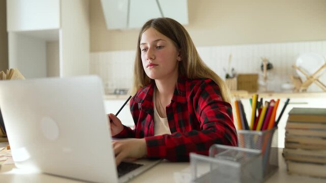 Girl studying and writing homework at desk with laptop and notebook focused concentration while using pencil for assignment, student working on schoolwork learning at home with bookstack and