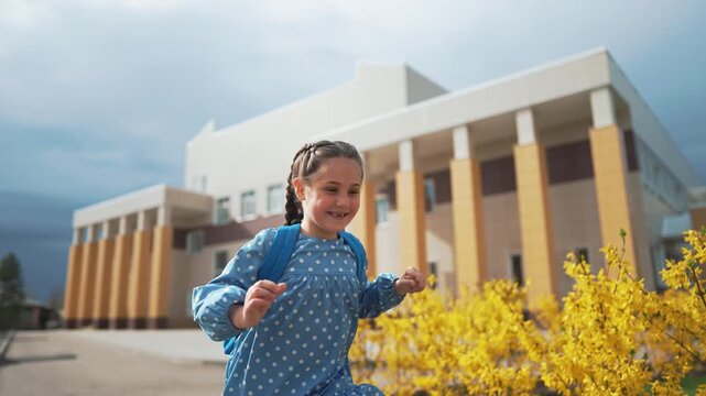 Girl raises arms celebrating after school child with backpack and polka dot dress smile near modern school building with yellow blossom, joyful girl shows play and joy in spring while leaving school
