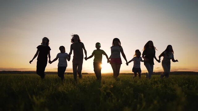 Group holding hands walking across grass field at sunset silhouetted family and friend figures move together child and adult linked in unity silhouette emphasizes connection togetherness and joy