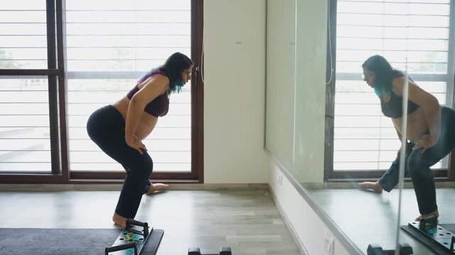 Indian female does leg stretching exercises in front of gym mirror in third trimester of pregnancy