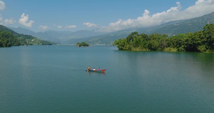 Aerial shot of tourists rowing a wooden colorful boat on calm blue Phewa Lake surrounded by small islands and dense forest in Pokhara Nepal.