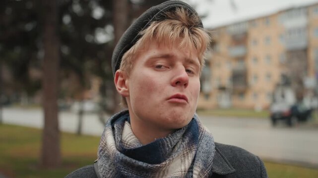 Young man smelling red rose on street with trees and apartments, contemplative closeup portrait, scarf and beret styling, soft bokeh background, cinematic intimate mood, creator admiring bloom