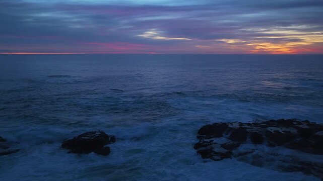 Stunning twilight seascape featuring waves crashing against dark rocky outcrops under dramatic purple and orange sunset sky. Calm ocean waters reflect vibrant colors as day turns to night