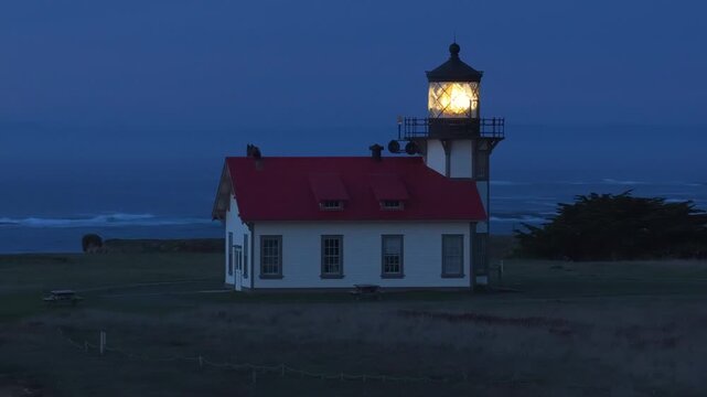Aerial view of Point Cabrillo Lighthouse glowing at night in Mendocino, California, USA. Historic white building with red roof stands on coastal grassland with Pacific Ocean waves in background