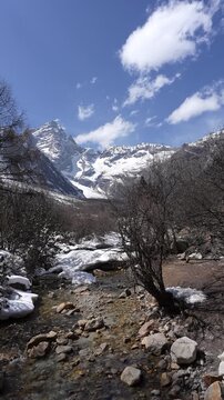 Snow-covered mountain valley with a clear rocky stream flowing through leafless trees under a bright blue sky, capturing a peaceful winter landscape in a remote natural wilderness setting.