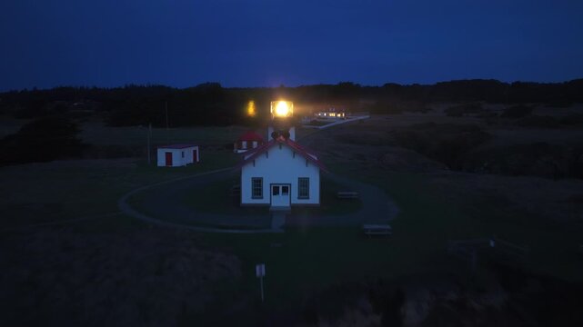 Night aerial footage of Point Cabrillo Lighthouse with its beacon glowing against dark blue sky. Waves crash on rocky coastline as the historic light station illuminates the Mendocino California coast