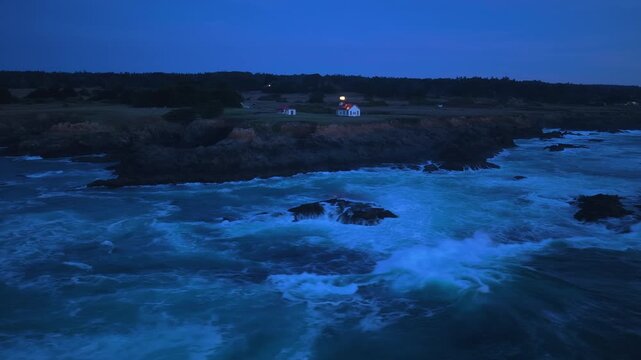 Night aerial view of Point Cabrillo Lighthouse in Mendocino, California, USA. Powerful ocean waves crash against rocky cliffs while the beacon shines brightly over the dramatic Pacific coastline