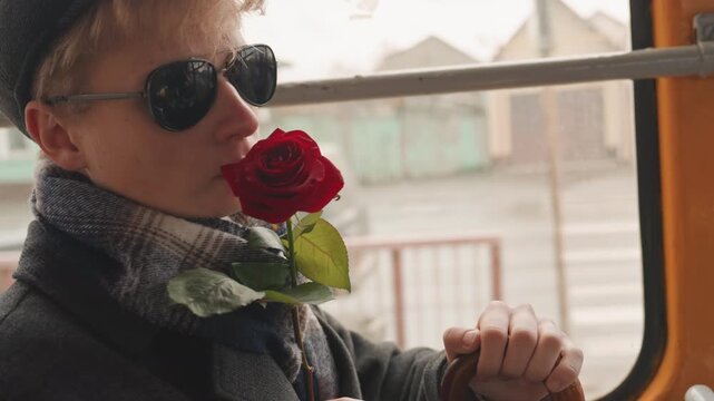 Profile white male creator wearing sunglasses and scarf with rose near lapel, looking through bus window at passing city, muted palette and cool cinematic mood, contemplative commuter scene