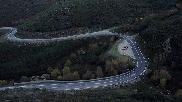 N338 mountain road with sharp hairpin bend and stone culvert bridge in Serra da Estrela, Central Portugal, showing parked vehicle, guardrails, shrub vegetation and steep valley terrain, drone orbits