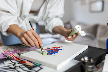Fototapeta premium Close up of female artist hand using a brush to apply light green acrylic paint onto an abstract canvas. Creative woman painter working on contemporary art in a bright modern studio with soft light.