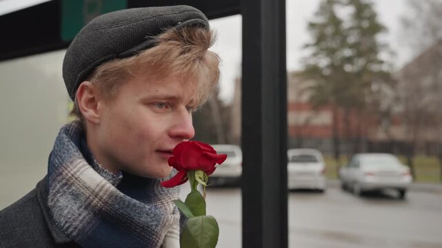 Young white man offering rose, creator rehearsing lines at bus shelter, expressive smile and gestures, scarf and cap, candid urban moment, playful audition vibe and quiet optimism