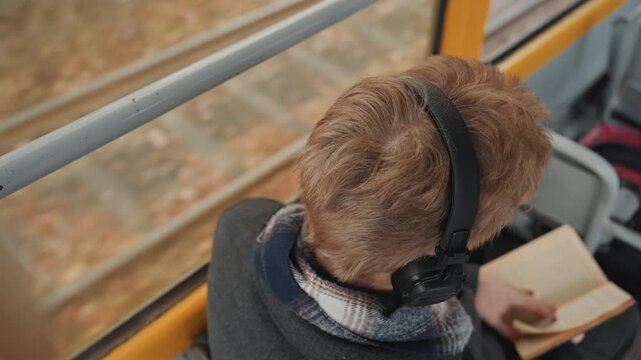 Young creator on bus reading notebook, wearing headphones by window, autumn tracks blurring outside, cozy scarf and seat, sketching ideas and jotting notes with quiet focus and reflective mood