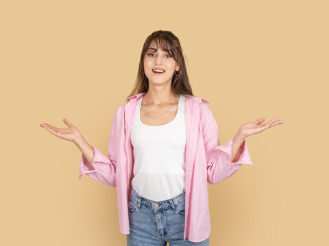 Smiling caucasian woman shrugging with open hands, expressing uncertainty or indifference concept idea image, wearing pink shirt and denim, isolated on beige background with copy space.