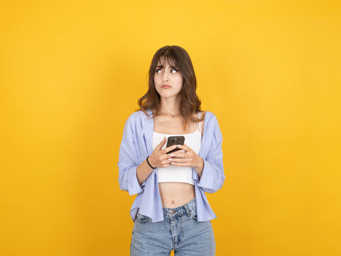 Unhappy caucasian woman holding smartphone and looking aside with doubtful expression, reacting to message or situation, wearing striped shirt, isolated on bright yellow background with copy space.