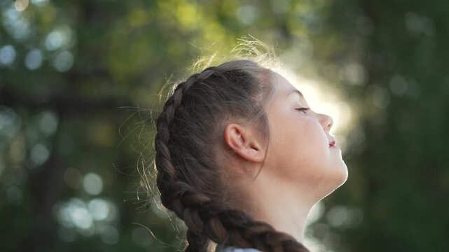Girl breathing slowly in warm sunlight, child with braided hair stands outdoors in nature, profile portrait shows closed eyes and calm expression, braid and hair halo catch light, sunlight on cheek