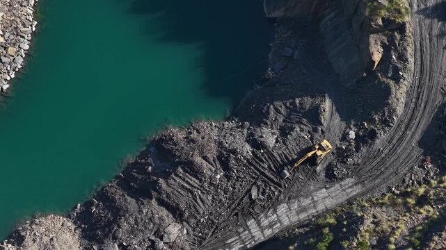 Aerial view of a yellow excavator stands in stark contrast to the dark earth of a quarry next to the bright turquoise water, Braga, Portugal.