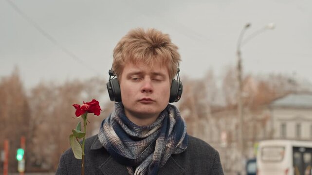 White creator with headphones flipping finger while holding rose, provocative artist making bold street statement, scarfed figure at bus stop, autumn city backdrop and playful confrontation captured