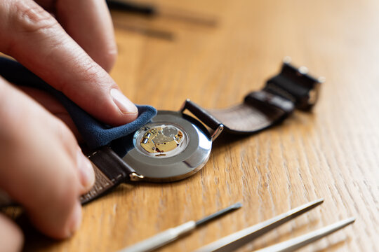 Hand is polishing the back of a wristwatch with a leather strap on a wooden table, with watchmaking tools visible in the scene