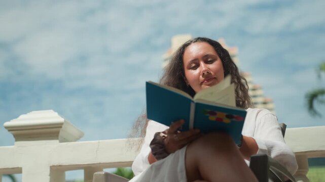 Smiling woman reading. Woman enjoying colorful book outdoors. Relaxed woman under bright sky reading colorful notebook. Woman smiling outdoors amidst tropical palms reading vibrant notebook