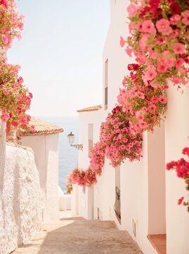 Colorful flowers in pink hues adorn whitewashed walls along a narrow street leading to the sea in a picturesque coastal village in Spain during summer