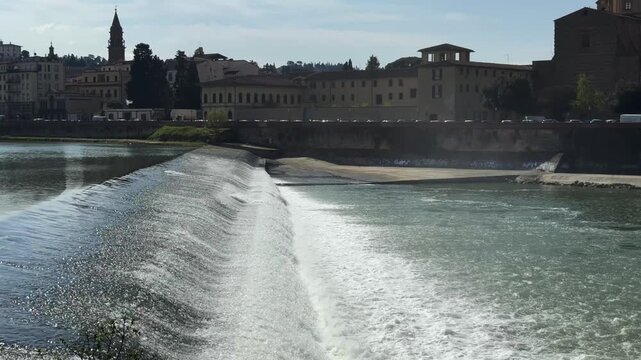 Water cascading over a low weir on the Arno River in Florence, with the historic riverside skyline and church tower visible in the background.