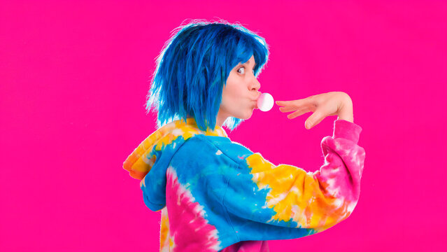 Profile of a young person with bright blue hair blowing a bubblegum bubble against a vibrant pink background
