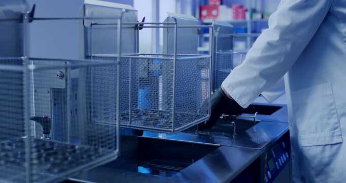Person in white lab coat placing metal wire basket with industrial parts into cleaning machine