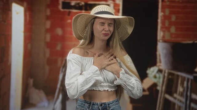 Young woman wearing a summer hat with bare midriff holds hands on chest in an unfinished building; pain vulnerability.