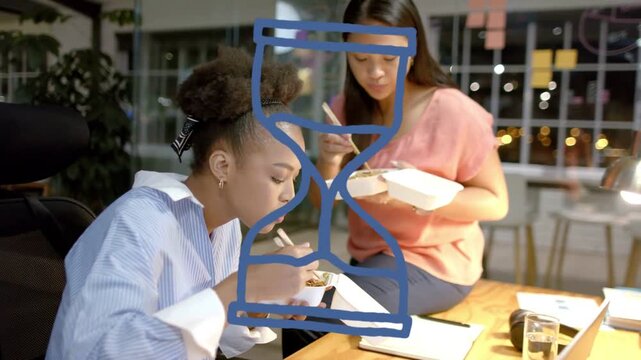 Two female coworkers in office working late, eating takeout, checking laptop while hourglass hiding
