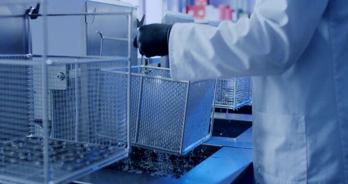 Technician in white lab coat lifting dripping metal basket from industrial cleaning machine