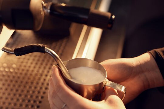 Barista frothing milk in metal pitcher with coffee machine wand in cafe, closeup