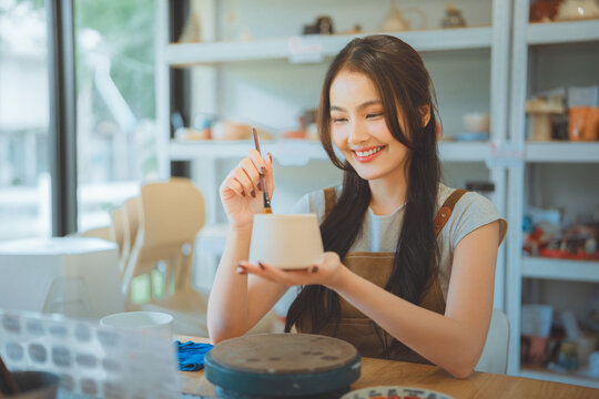 Happy young Asian woman artisan smiling while painting a ceramic pot with a brush in a pottery studio workshop; a creative mindful hobby, art therapy, and DIY lifestyle concept for small business