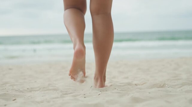 Barefoot female legs walking toward ocean on island shore, contemplative shoreline scene with gentle waves and horizon, cinematic overcast light, reflective mood of beach wanderer.