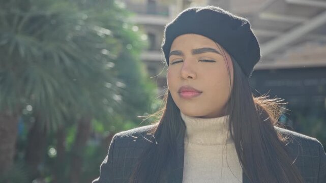 Young hispanic woman in a black beret and cream turtleneck gazes straight with neutral expression on a street; urban poise.