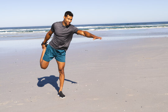 Man stretching and balancing on leg at sandy beach with teal shorts, running shoes, fitness tracker