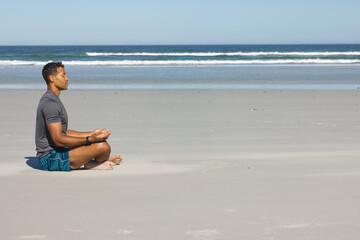 Fototapeta premium Mid-adult man meditating cross-legged on beach in sportswear with smartwatch, copy space