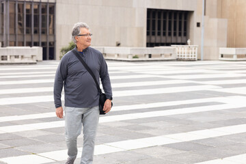 Senior man walking across striped plaza by concrete building with black crossbody bag, copy space © wavebreak3