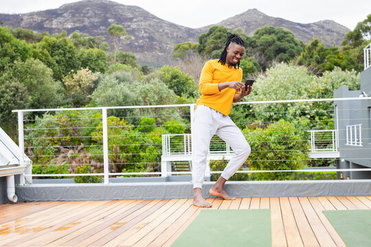 African American man standing barefoot on rooftop deck holding phone near two yoga mats, copy space