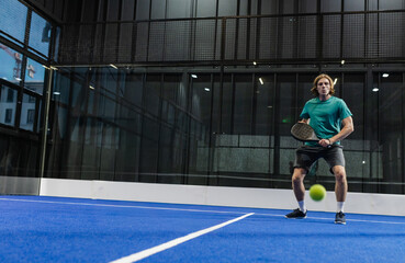 Obraz premium Man standing right of padel court in teal sportswear holding racket watching green ball, copy space