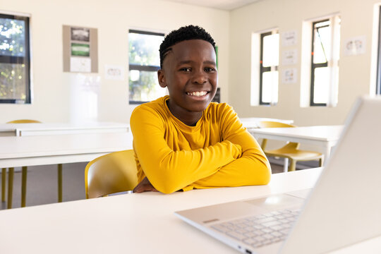 African teen in yellow longsleeve shirt smiling sitting at classroom table using open silver laptop