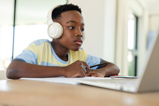 African American child sitting at wooden home desk wearing headset holding pen looking at laptop