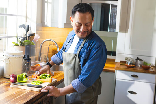 Mature Asian man in apron chopping pepper and red onion on wooden board in home kitchen