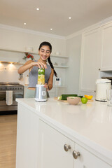 Adult Indian woman preparing green smoothie at kitchen island in grey tank top, using blender jar © wavebreak3