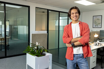 Fototapeta premium Asian man wearing red shirt and jeans standing in open-plan office near white planter, copy space