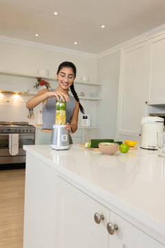 Adult Indian woman preparing green smoothie at kitchen island in grey tank top, using blender jar