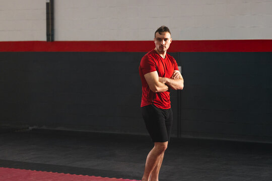 Man standing cross-armed on mat in gym by red stripe pipe, red shirt barefoot, copy space