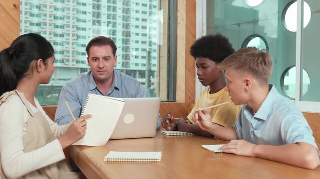 Smart girl ask teacher a question while show idea while diverse student working classwork. Group of multicultural children pointing at notebook while happy boy sharing idea at classroom. Edification