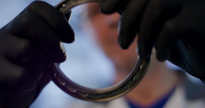 Hands in black gloves holding and inspecting a steel ball bearing in a laboratory or industrial setting