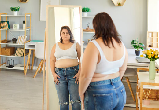 Photo of obese fat woman dressed in white top and jeans, looking at her reflection in mirror, doesnt like her body, has access weight, gaining to be fit, needs to keep diet. Obesity, health problems